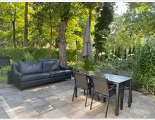 Terrasse mit Steinboden, einem schwarzen Ledersofa, einem schwarzen Esstisch mit vier Stühlen und einem geschlossenen grauen Sonnenschirm; im Hintergrund Bäume, Büsche und blühende Pflanzen in einem grünen Garten.