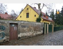 Straßenszene mit einem gelben Haus mit Spitzdach und roten Ziegeldachziegeln, eingefasst von einer alten, mit Graffiti verzierten Mauer. Der Boden besteht aus Kopfsteinpflaster, und im Hintergrund sind Bäume und ein Zaun sichtbar.