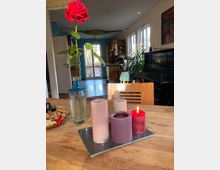 Ein Esszimmer mit einem Holztisch im Vordergrund, auf dem eine Vase mit einer roten Rose und mehrere farbige Kerzen auf einem Tablett stehen, eine davon brennend. Im Hintergrund eine offene Küche mit Holzschränken, sowie ein schwarzes Klavier an der rechten Wand.