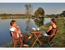 Zwei Personen sitzen auf Holzstühlen an einem kleinen Tisch mit Getränken und Essen, direkt an einem ruhigen See oder Fluss. Im Hintergrund sind Bäume, Wiesen und ein Auto zu sehen, das Ufer spiegelt sich im Wasser.
