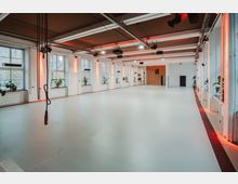 Großer, offener Raum mit weißen Wänden, poliertem hellen Boden und einer Decke mit sichtbaren Trägern und Beleuchtungselementen. Entlang der Fensterreihe befinden sich Pflanzen auf Fensterbänken, und im Hintergrund ist eine Holzwand mit einer Tür sichtbar.