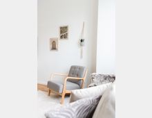 Minimalistisch eingerichtetes Wohnzimmer mit einem grauen Sessel mit hell hölzernem Rahmen auf hellem Teppichboden. An der weißen Wand hängen zwei gerahmte Bilder und ein kleiner Pflanzenhalter aus Makramee.