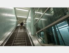 Ein moderner U-Bahn-Bereich mit einer Rolltreppe, die nach oben führt. Links sind hellgrüne, geflieste Wände, während rechts Glasfenster einen Blick auf technische Strukturen im Inneren erlauben.