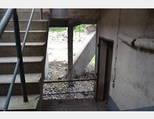 Eine Betoninnentreppe mit Metallgeländer führt in einen Bereich mit sichtbarem Bauschutt und freiliegender Stahlarmierung. Rechts ist eine alte, beschädigte Wand mit freiliegenden Rohren, die ebenfalls Abnutzungserscheinungen zeigt.