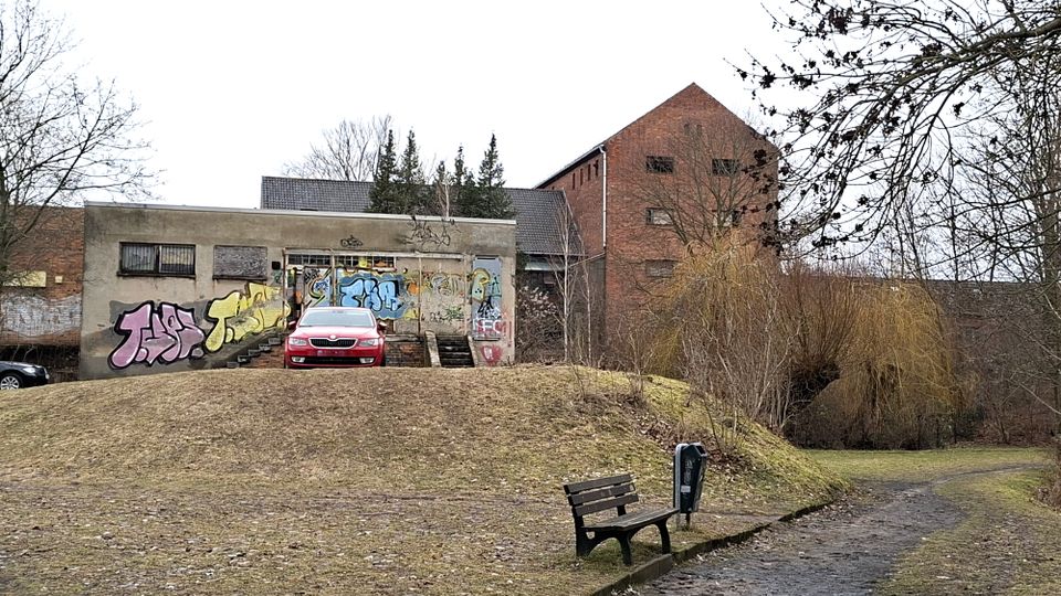 Blick auf ein niedriges, graues Gebäude mit Graffiti und Treppenaufgang, daneben ein höherer Backsteinbau. Davor liegt eine grasige Böschung mit Parkbank und einem Weg, umgeben von kahlen Bäumen und Sträuchern.