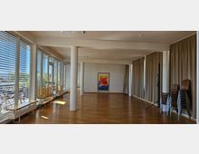 Heller Veranstaltungsraum mit Parkettboden, großen Fenstern mit Jalousien auf der linken Seite und beige Vorhängen rechts. Stapelstühle stehen in der Ecke, zusammen mit einem zentral aufgehängten Farbbild an der weißen Wand.