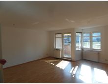 Helles Wohnzimmer mit Laminatboden, großen Fenstern und direktem Zugang zu einem Balkon. Heizkörper befinden sich unter den Fenstern, und draußen ist ein gepflegter Außenbereich sichtbar.