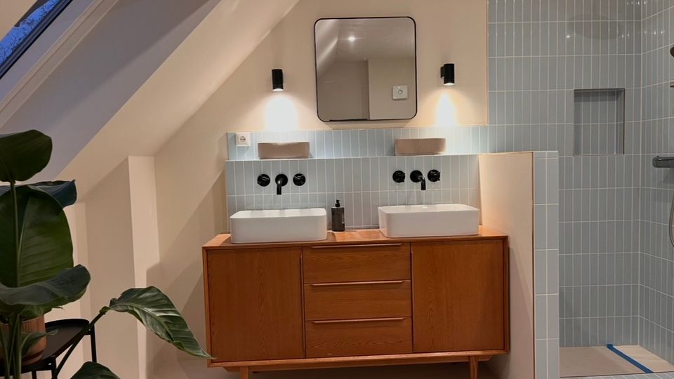 Modernes Badezimmer in einem Dachgeschoss mit Holz-Waschtisch, zwei rechteckigen Waschbecken und blauen Wandfliesen. Eine Glasdusche mit Regenduschkopf befindet sich rechts, und links steht eine große, grüne Pflanze auf einem schwarzen Gestell.
