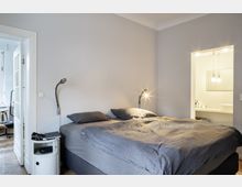 Schlafzimmer in modernem Design mit einem großen Doppelbett, grauer Bettwäsche und zwei Wandleselampen. Im Hintergrund ist eine offene Tür, die zu einem Tageslichtbad mit hellem Waschbereich führt.