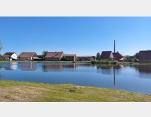 Dorfansicht mit mehreren Bauernhäusern und Wirtschaftsgebäuden am Ufer eines Teichs, im Hintergrund ein hoher Schornstein. Der Himmel ist klar und blau, während die Landschaft sich im Wasser spiegelt.
