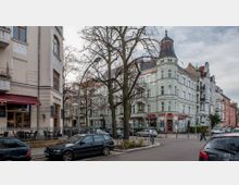 Straßenszene mit mehrstöckigen Altbaugebäuden im historischen Stil, darunter ein Eckhaus mit hellem Putz und verzierten Fassaden. Am unteren Rand befinden sich Geschäfte, ein Café mit rotem Markisendach, parkende Autos und spärlich belaubte Bäume.