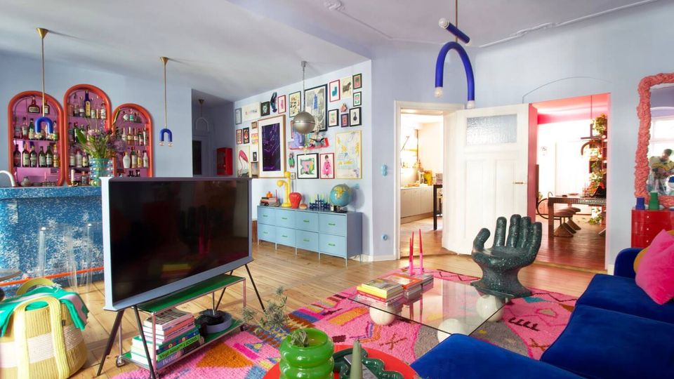Eclectic, colorful living room with light wood floors, pale blue walls, and a large blue sofa around a glass coffee table on a bright patterned rug. The space features a gallery wall of framed art, arched shelving filled with bottles, modern pendant lights, and open double doors leading to a hallway and another room.
