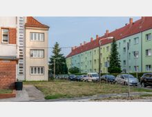 Wohnsiedlung mit mehrstöckigen Wohnhäusern in pastellfarbenem Putz (grün und beige) und roten Ziegeldächern. Im Vordergrund geparkte Autos, Gehweg und eine Grünfläche, umgeben von Straßenlaternen und Verkehrsschildern.