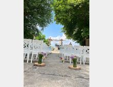 Ein Außenbereich mit einer Zeremonie-Setup: Weiße Stühle sind in Reihen angeordnet, entlang eines Weges mit Blumen in Glasvasen dekoriert. Im Hintergrund ist ein hölzerner Bogen unter Bäumen sichtbar, dahinter ein klarer Himmel und ein Gebäude in der Ferne.