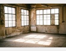 Leerer, heruntergekommener Industrieraum mit großen Sprossenfenstern, abblätterndem Putz und sichtbaren Leitungen an den Wänden. Betonboden mit Sonnenlichtreflexen, Heizkörpern unter den Fenstern und einfachen Leuchtstofflampen an der Decke.