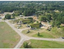 Luftaufnahme eines ehemaligen Militärflugplatzes mit mehreren grasbewachsenen Bunkern, alten Kampfflugzeugen und einer asphaltierten Rollbahn, umgeben von Waldgebieten. Im Hintergrund sind Baumreihen und einige Gebäude zu erkennen.