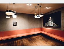 Ein moderner Warteraum mit einer langen, orangefarbenen Eckbank aus gepolsterten Sitzflächen vor grauen Betonwänden. An der Wand hängen zwei Kunstwerke, ein kleines Bild und ein großes, schwarzes Gemälde mit einer Collage; über der Szene hängen hängende Designlampen.