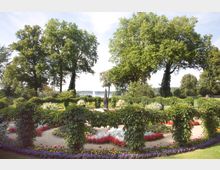 Ein sorgfältig gestalteter Garten mit kreisförmigen Blumenbeeten in leuchtenden Farben, umgeben von grünen Rankgittern. Im Hintergrund stehen große, üppig belaubte Bäume, und ein Brunnen mit einer Wasserfontäne ist in der Ferne zu sehen.