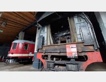 Innenansicht eines historischen Lokschuppens mit einer großen Dampflok-Bedienfläche im Vordergrund und einem rot-beigen Schienenfahrzeug im Hintergrund. Die Decke besteht aus sichtbaren Holzbalken, und die Beschilderung liefert technische Informationen.