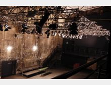 Innenansicht eines industriellen Veranstaltungsraums mit rauen, teils beschädigten Wänden und einer Metalltreppe auf der rechten Seite. An der Decke hängt ein komplexes Gerüst mit zahlreichen Scheinwerfern und Beleuchtungselementen.