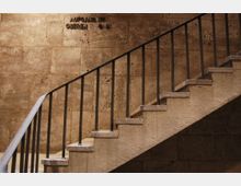 Treppe mit Metallgeländer vor einer Wand aus beigefarbenen Steinplatten; darüber befindet sich eine eingravierte Beschriftung mit der Aufschrift "Aufgang zum oberen L. 6.-8.".