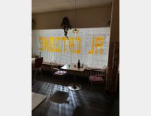 Innenraum eines Cafés mit gläserner Fensterfront, auf der in gelber Schrift „AL CATZONE“ zu lesen ist. Vor dem Fenster steht ein Tisch mit zwei Stühlen aus Metall, dekoriert mit einer Weinflasche, Kerzenhaltern und kleinen Gegenständen; an der Decke hängt eine schlichte Lampe.