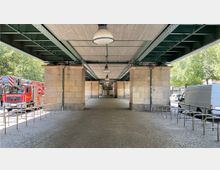 Unterführung einer Stahlbeton-Brücke mit grünen Stahlträgern und eingelassenen Deckenlampen. Seitlich befinden sich massive Steinpfeiler, dahinter sind Fahrradbügel sowie parkende Fahrzeuge und Bäume zu erkennen.