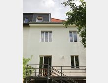 Zweistöckiges Wohnhaus mit weißer Fassade, einem verglasten Balkon im Dachgeschoss und einer kleinen Terrasse mit Metallgeländer im Erdgeschoss. Im Vordergrund stehen Bäume und Büsche.