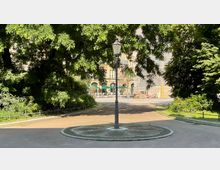 Kreisförmiger Platz mit Kopfsteinpflaster und einer Straßenlaterne in der Mitte, umgeben von Gehwegen, dichten Bäumen und Sträuchern; im Hintergrund ist eine belebte Straße mit einem historischen Gebäude und grünen Sonnenschirmen zu sehen.