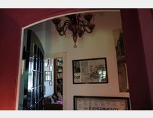 Innenraum mit einem roten Türbogen, einer geöffneten schwarzen Glastür und einer weißen Wand, die mit gerahmten Postern dekoriert ist. Im Hintergrund ist ein weiterer Raum mit Bücherregalen, Bildern und einem bunten Sofa sichtbar; zudem hängt von der Decke ein Kronleuchter.