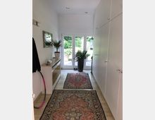 Ein heller Flur mit beigen Fliesen, weißen Wänden und einer durchgehenden Einbauschrankwand rechts. Vor den Fenstertüren, die zu einem Garten führen, stehen Pflanzen in Töpfen, und auf dem Boden liegen orientale Teppiche.