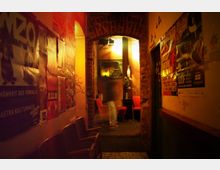 Schmaler, gelblich beleuchteter Flur in einer Bar oder einem Veranstaltungsort mit Postern an den Wänden und roten Polsterstühlen am Ende des Korridors. Das Interieur ist von Backsteinbögen und einer warmen, gemütlichen Atmosphäre geprägt.
