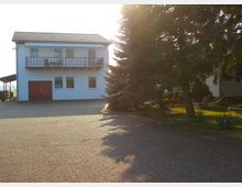 Weiß gestrichenes zweistöckiges Wohnhaus mit Balkon, roten Türen und Ziegeldach, umgeben von einem gepflasterten Vorhof und großen Nadelbäumen im Sonnenlicht.