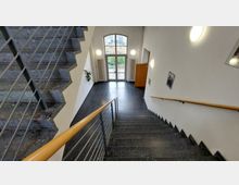 Treppenhaus mit grauen Steinfliesen und einem Geländer aus Metall mit Holzhandlauf. Im Hintergrund eine gläserne Doppeltür, die zu einem Außenbereich mit Pflanzen führt; die Wände sind weiß, und es sind runde Wandlampen sowie Bilder sichtbar.