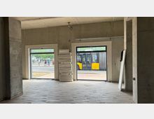 Innenansicht eines unfertigen Gebäudes mit Betonwänden und Fliesenboden. Zwei große Fenster bieten Blick auf eine Bushaltestelle mit stehenden Personen und einem gelben Bus draußen; Styroporplatten sind neben einem Fenster gestapelt.