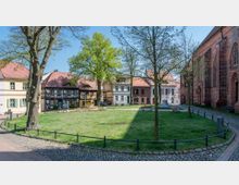 Kleiner Platz mit Rasenfläche, umgeben von historischen Fachwerkhäusern und einem Gebäude aus rotem Backstein, das an eine Kirche erinnert. Im Vordergrund verlaufen gepflasterte Wege, und der Platz ist mit Bäumen und schmiedeeisernen Geländern gestaltet.