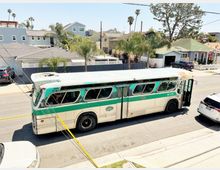 Ein alter, türkis-weiß lackierter Bus mit abgenutztem Äußeren steht auf einer Wohnstraße, umgeben von Palmwerk und niedrigen Häusern mit Garagen und Vorgärten. Im Hintergrund sind zusätzliche Fahrzeuge und moderne Wohnhäuser zu sehen.