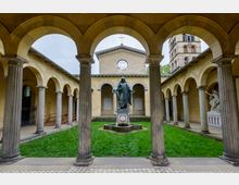 Offener Kreuzgang mit Rundbögen und Säulen um einen zentralen Grasbereich mit einer Statue im Mittelpunkt, dahinter eine kleine Kapelle mit Rundfenster; seitlich ist ein Kirchturm zu sehen.
