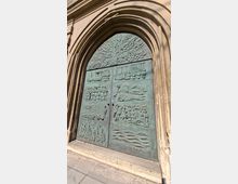 Großes, grün patiniertes Bronzetor mit Reliefdarstellungen, eingefasst von einem hohen, gestuften Spitzbogenportal aus hellem Stein. Das Portal liegt an der Außenfassade eines historischen Gebäudes und steht auf einer flachen Steinstufe mit gepflastertem Boden davor.