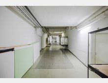 Langer, weiß gestrichener Flur mit sichtbaren Rohren an der Decke und Vitrinen entlang der Wände. Der Boden besteht aus glatten, grauen und beigen Flächen, und am Ende des Gangs ist ein weiterer Durchgang sichtbar.