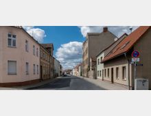 Straßenansicht einer ruhigen Wohnstraße mit zweigeschossigen Häusern, meist in hellen Farben verputzt oder verklinkert, und Satteldächern. Der Himmel ist teils bewölkt, und am Straßenrand stehen vereinzelt parkende Autos.
