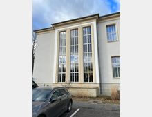 Ein modernes Gebäude mit heller Fassade, großen vertikalen Fenstern und beigefarbenen Fensterrahmen. Im Vordergrund steht ein graues Auto auf einem Parkplatz, umgeben von einigen Pflanzen und kahlen Sträuchern.