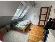 Kleines Dachgeschoss-Schlafzimmer mit schrägen Wänden, zwei großen Dachfenstern und Holzboden. Im Raum stehen ein niedriges Bett, ein weißer Kleiderschrank und eine Holztür mit Glaseinsätzen.
