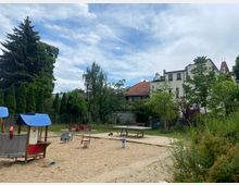 Ein kleiner Spielplatz mit einem Sandbereich, der von Bäumen und Sträuchern umgeben ist. Spielgeräte, darunter ein Spielhäuschen mit blauem Dach und Wippen, befinden sich im Vordergrund, während im Hintergrund Wohnhäuser mit roten Ziegeldächern zu sehen sind.