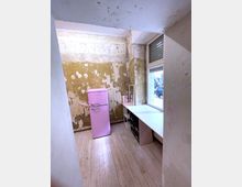 Ein kleiner Raum mit abgenutzten Wänden und einem hellen Holzfußboden. Ein rosa Kühlschrank steht an der linken Wand, daneben ein weißer Schreibtisch mit Regalfächern, vor einem Fenster mit Blick nach draußen.