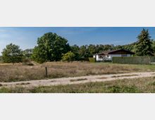 Ein eingeschossiges Wohnhaus mit weißer Fassade und dunklem Satteldach steht in einer ländlichen Umgebung mit Grasflächen, einem Feldweg und großen Bäumen im Hintergrund. Eine Hecke und ein Zaun umgeben das Grundstück.