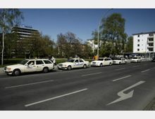 Reihe von cremefarbenen Taxis, die entlang einer Straße parken, umgeben von Grünflächen, Bäumen und mehrstöckigen Gebäuden. Im Hintergrund ist ein Bürogebäude mit einem "Hochtief"-Schild sichtbar.