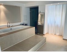 Modernes Badezimmer mit einer freistehenden Badewanne auf einer erhöhten Podestkonstruktion aus beigen Fliesen, einem großen Spiegel, einer Glasdusche im Hintergrund und bodenlangen weißen Vorhängen auf der rechten Seite.