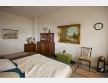 Ein Schlafzimmer mit einfacher Einrichtung, bestehend aus einem Doppelbett mit weißer Bettdecke im Vordergrund, antiken Holzmöbeln wie einem Schrank und einem Sideboard sowie Dekorationselementen wie zwei gerahmten Gemälden an der Wand, einer Wanduhr und einem Stuhl. Der Raum hat helle Wände, einen schlichten Bodenbelag und ist mit Teppichen ausgelegt.