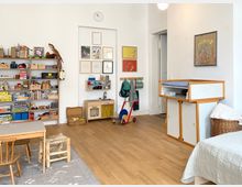 Heller, weiß gestrichener Spiel- und Wohnraum mit Holzfußboden, niedrigem Tisch und kleinen Stühlen sowie offenen Regalen voller Spielzeug und Bücher. An der Wand hängen mehrere gerahmte Bilder über einer Kinder-Spielküche; daneben Garderobenhaken und ein offener Durchgang zu einem Flur, rechts ein Wickel- oder Aufbewahrungsmöbel.